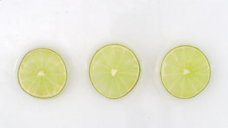 Wasserspritzer-In-Zeitlupe-Auf-Drei-Scheiben-Grüner-Limette,-Die-Auf-Weißem-Hintergrund-Im-Wasser-Liegen